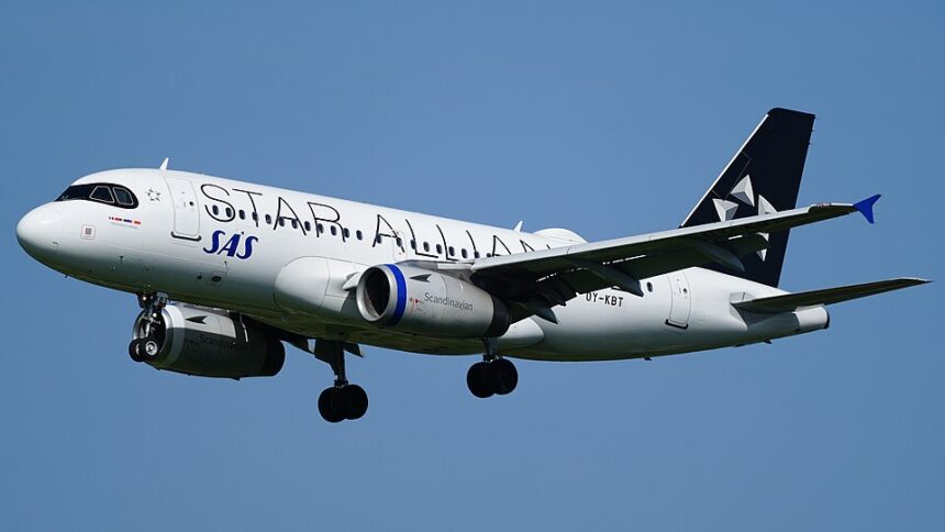 Side view of a Scandinavian Airlines (SAS) Airbus A320 in flight against a clear blue sky, captured on final approach with its landing gear fully extended. The white fuselage features “Scandinavian” written in large grey lettering along the side, with the blue “SAS” logo near the front. The engines beneath the wings are painted dark blue, and the tail is also dark blue with a white star‑shaped emblem. The aircraft’s wings are slightly flexed, with flaps deployed, and the nose is angled slightly downward as it descends.