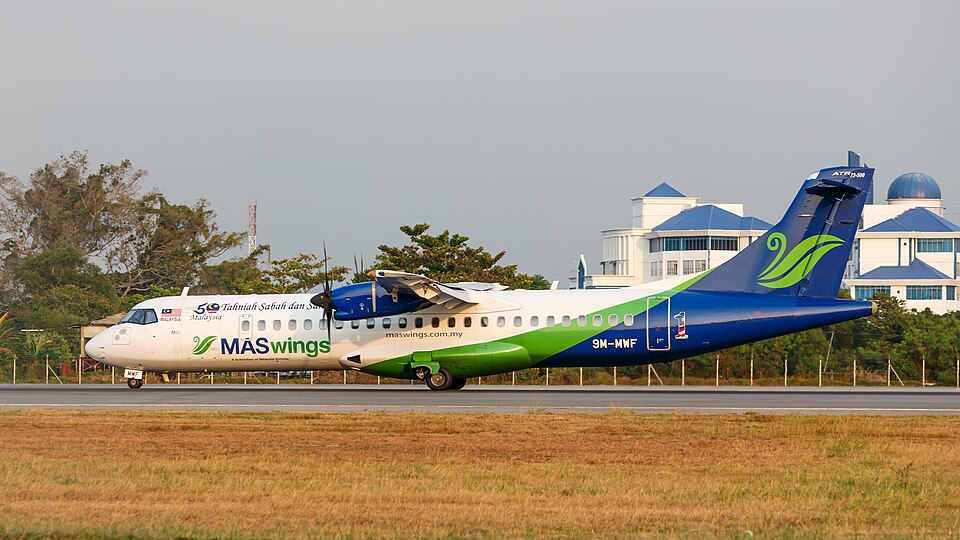 A MASwings ATR 72-500 turboprop aircraft, registered 9M-MWF, taxis along the runway at Kota Kinabalu International Airport in Sabah, Malaysia. The aircraft is painted in the airline’s blue, green and white livery, with airport buildings and trees visible in the background under clear daylight conditions.