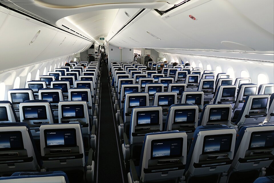 A forward-facing view of an empty Lufthansa economy class airplane cabin. Rows of modern passenger seats with dark blue upholstery flank a single central aisle.
