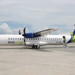 An ATR 72-500 twin-engine turboprop passenger airliner, operated by MASwings with tail number 9M-MWA, sits parked on the airport tarmac on a bright, partly cloudy day.