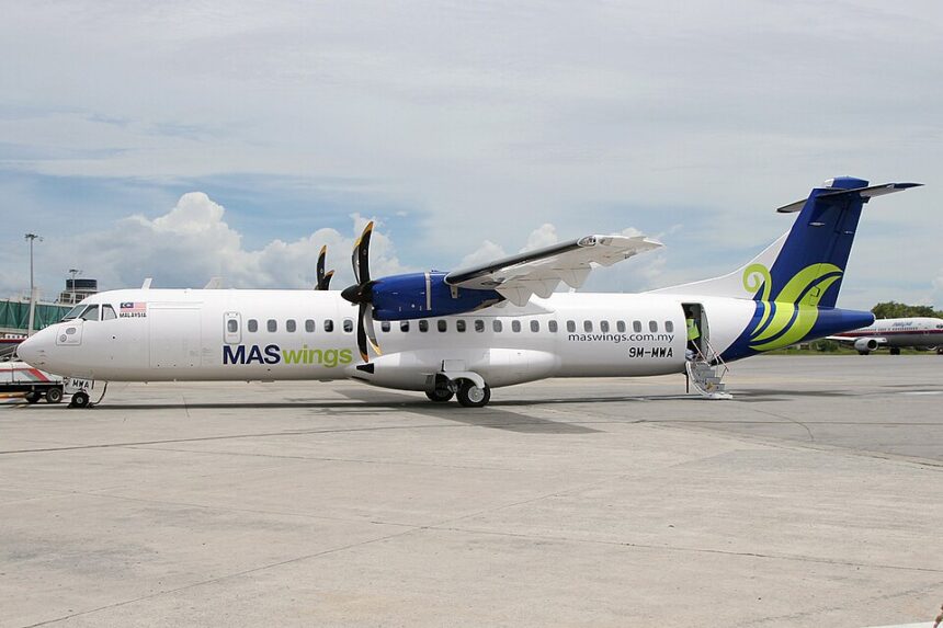 An ATR 72-500 twin-engine turboprop passenger airliner, operated by MASwings with tail number 9M-MWA, sits parked on the airport tarmac on a bright, partly cloudy day.