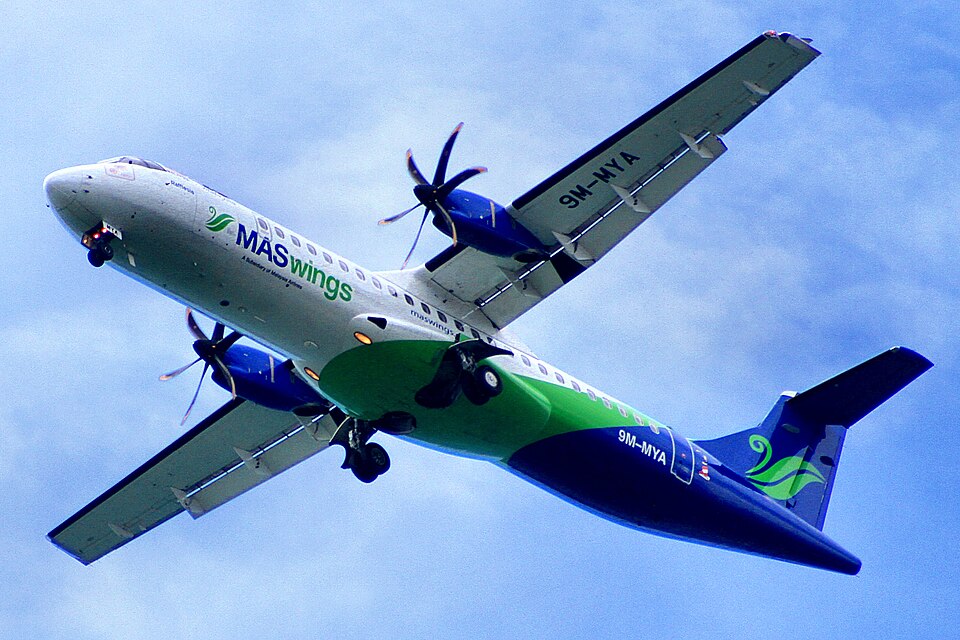 A low-angle, full shot captures a white, green, and blue MASwings twin-engine turboprop airplane soaring diagonally upward toward the upper right.