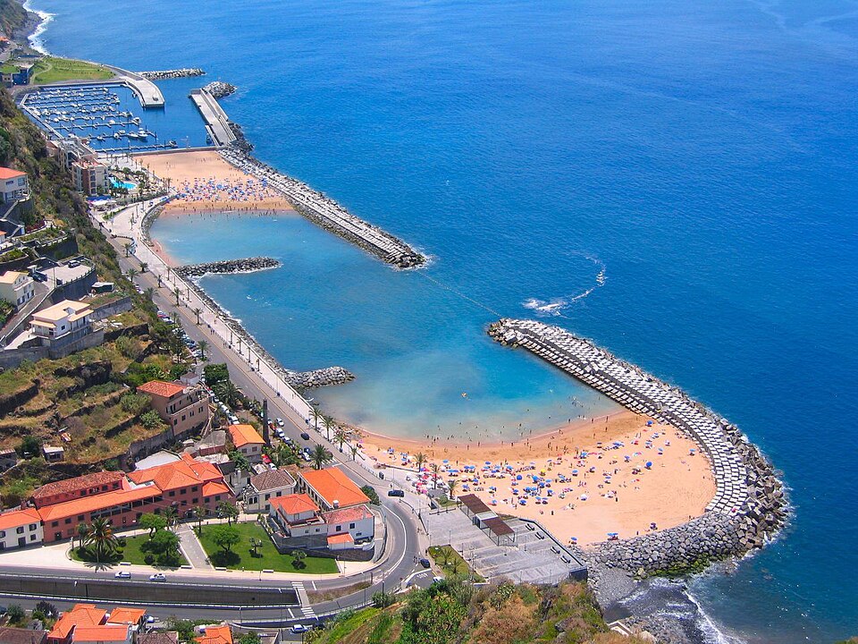 Aerial view of Calheta Beach in Madeira. The water is both light and royal blue. The sand is filled with chairs and umbrellas for those who are enjoying a day at the beach.
