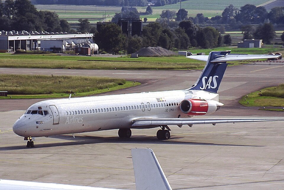 A Scandinavian Airlines (SAS) MD-80 series aircraft taxiing on an airport tarmac, shown in profile view. The silver and white plane displays "Airlines" text on its fuselage with the registration "SE-DMA" near the rear. Its distinctive blue tail fin prominently features the white "SAS" logo, and the rear-mounted engine has a red cowling with "Scandinavian" branding. In the foreground, the wing tip of another aircraft is visible. The background reveals a rural airport setting with hangar buildings, storage tanks, a gravel pile, and lush green fields and trees extending into the distance.