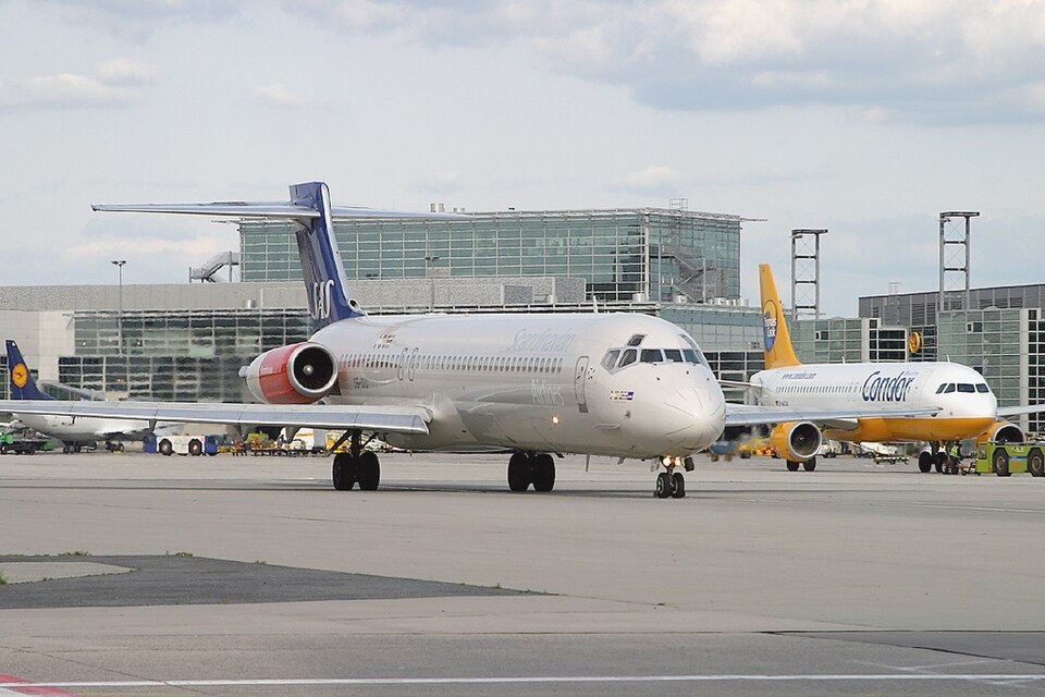A Scandinavian Airlines (SAS) MD-80 series aircraft taxiing on the tarmac at an airport, viewed from a near front-on angle. The silver and white plane displays the "Scandinavian" name along its fuselage and has its characteristic rear-mounted engines visible. To the right, a Condor airliner with a distinctive yellow and white livery is parked at the terminal gate. On the far left, a partial view of a Lufthansa aircraft tail can be seen. The modern glass-fronted terminal building stretches across the background, with ground service vehicles and equipment scattered around the apron under a partly cloudy sky.