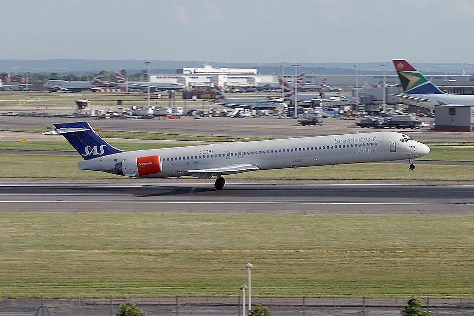 A Scandinavian Airlines (SAS) McDonnell Douglas MD-82 aircraft taking off from a runway. The long, narrow plane is captured mid-rotation with its nose wheels off the ground. It features a white fuselage with "Scandinavian Airlines" text, distinctive red rear-mounted engines, and a blue tail fin with the white SAS logo. In the background, other commercial airplanes and airport infrastructure are visible under a clear, bright sky.