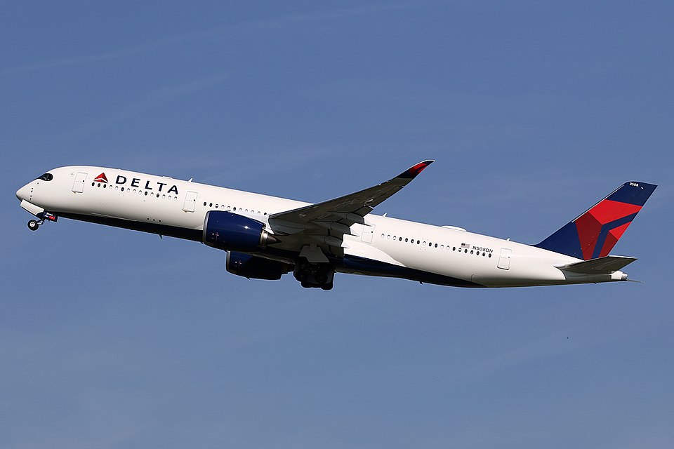 A Delta Air Lines Airbus A350 wide-body jet airliner climbing steeply into a clear blue sky, with its white fuselage, dark blue engines, and distinctive red-and-blue tail fin prominently visible. The aircraft’s nose landing gear is in the process of retracting.