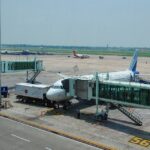 A high-angle, daylight shot captures an IndiGo commercial airplane parked on the tarmac at Kolkata Airport. The plane is docked with a green-tinted glass passenger jet bridge at Gate 56.