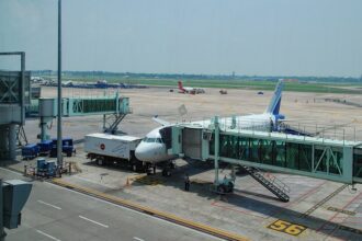 A high-angle, daylight shot captures an IndiGo commercial airplane parked on the tarmac at Kolkata Airport. The plane is docked with a green-tinted glass passenger jet bridge at Gate 56.