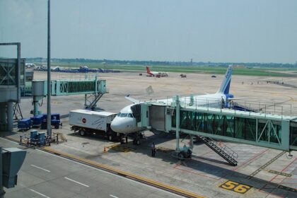 A high-angle, daylight shot captures an IndiGo commercial airplane parked on the tarmac at Kolkata Airport. The plane is docked with a green-tinted glass passenger jet bridge at Gate 56.
