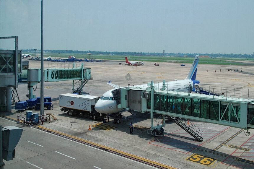 A high-angle, daylight shot captures an IndiGo commercial airplane parked on the tarmac at Kolkata Airport. The plane is docked with a green-tinted glass passenger jet bridge at Gate 56.