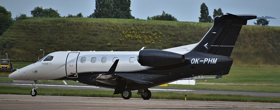 View of an Embraer Phenom 300 business jet parked on an airport apron. The aircraft features a distinctive black-and-white paint scheme, with a white nose and fuselage.