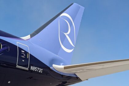A close-up view of the tail section of a Riyadh Air Boeing 787 aircraft against a clear blue sky. The livery features a gradient from dark indigo purple on the fuselage to light periwinkle blue on the vertical stabilizer, which displays the airline's distinctive white stylized "R" logo. The registration number "N8572C" is visible on the rear fuselage near a service door, and the aircraft's horizontal stabiliser extends into the frame.