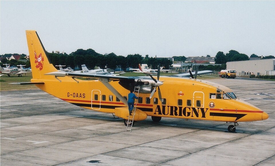 A vintage image of a Short 360 Aurigny on the ground. The aircraft is yellow, and it is parked on a concrete floor. The sky above it is mostly grey and on the side of the aircraft there's a man on a ladder.