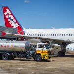 A TAM Airlines aircraft parked on an airport tarmac, with a silver Shell fuel tanker truck positioned beside it for refueling and a cargo loader servicing the rear of the fuselage.