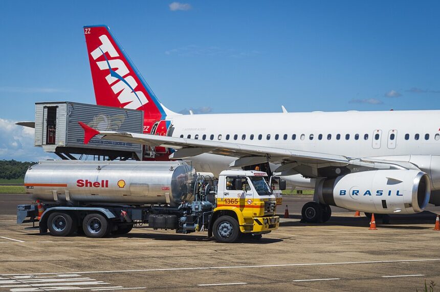 A TAM Airlines aircraft parked on an airport tarmac, with a silver Shell fuel tanker truck positioned beside it for refueling and a cargo loader servicing the rear of the fuselage.