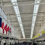 The interior of a large, modern airport terminal check-in hall with a high, white steel truss ceiling and skylights. Rows of check-in counters with yellow zone signs (5E, 5F) line the right side, where travellers are queued. On the left, large windows illuminate international flags, including Swiss and Turkish flags, hanging from an upper level.