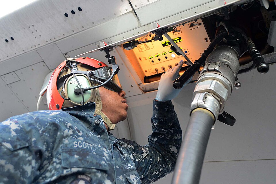 A U.S. Navy service member wearing a blue digital camouflage uniform with the name tape "SCOTT," a red cranial helmet, and ear protection, operating controls inside an open refueling panel on the underside of an aircraft. A large fuel hose with a metal nozzle is connected to the aircraft, and the illuminated control panel displays various gauges and indicators.