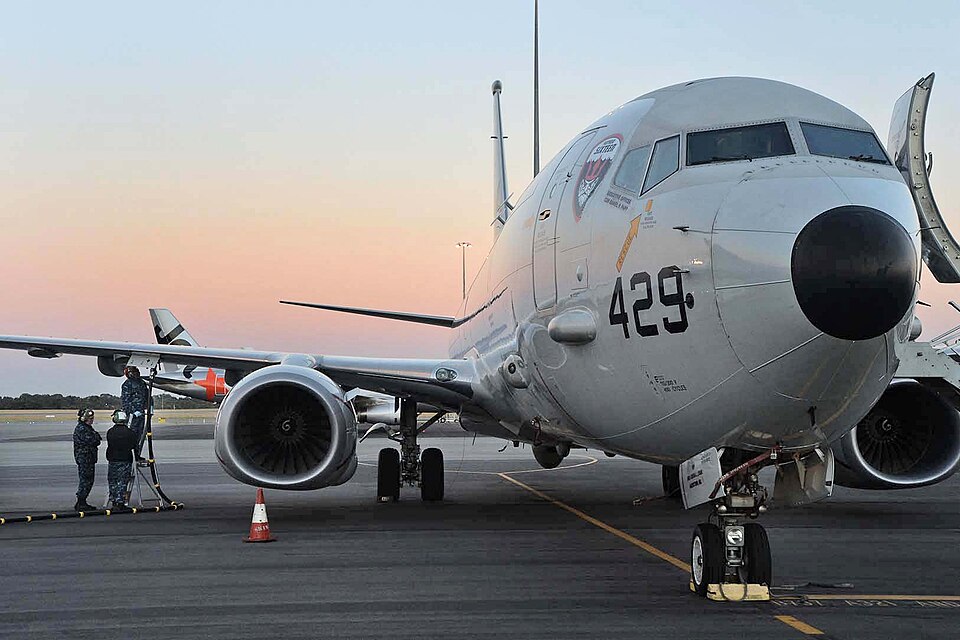 A U.S. Navy Boeing P-8A Poseidon maritime patrol aircraft, bearing the side number "429," parked on a tarmac during twilight. Two crew members in blue working uniforms are positioned near the left wing; one stands on a maintenance ladder attending to the port engine while the other assists from below. The aircraft's grey fuselage features squadron insignia near the cockpit, and an orange safety cone is placed on the ground beside the engine.
