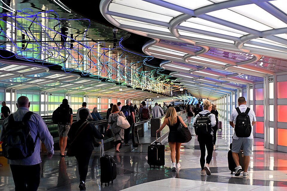 Travellers with rolling luggage walk through the underground pedestrian tunnel at Chicago O'Hare International Airport, featuring the colourful neon light installation Sky's the Limit suspended from the ceiling above moving walkways, with illuminated rainbow-tinted glass block walls lining the corridor.