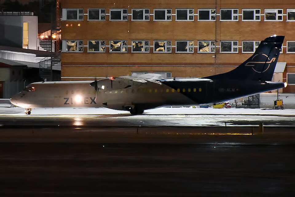 A side-profile, nighttime photograph of a Zimex Aviation ATR 72-202(F) twin-engine turboprop cargo aircraft parked on a wet, brightly illuminated tarmac.