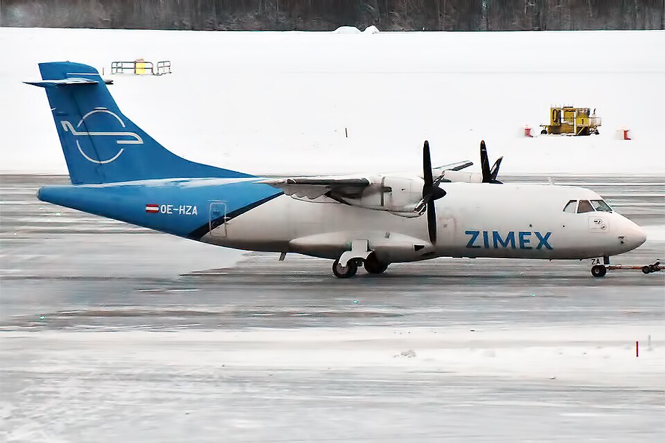 A side-profile photograph of a Zimex Aviation ATR twin-engine turboprop aircraft parked on a slushy, wet tarmac during winter.
