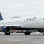 A Delta Air Lines Boeing 767 taxiing on a wet airport tarmac with patches of snow on the ground, viewed from the front-right angle. The aircraft features Delta's red, white, and blue livery with the prominent "DELTA" titles and widget logo on the fuselage, and a ground vehicle is visible near the nose gear under an overcast sky.