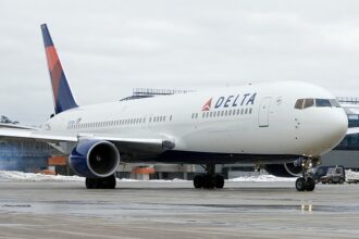 A Delta Air Lines Boeing 767 taxiing on a wet airport tarmac with patches of snow on the ground, viewed from the front-right angle. The aircraft features Delta's red, white, and blue livery with the prominent "DELTA" titles and widget logo on the fuselage, and a ground vehicle is visible near the nose gear under an overcast sky.