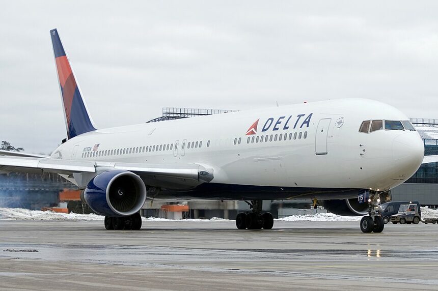 A Delta Air Lines Boeing 767 taxiing on a wet airport tarmac with patches of snow on the ground, viewed from the front-right angle. The aircraft features Delta's red, white, and blue livery with the prominent "DELTA" titles and widget logo on the fuselage, and a ground vehicle is visible near the nose gear under an overcast sky.