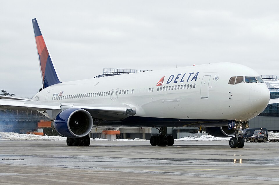 A Delta Air Lines Boeing 767 taxiing on a wet airport tarmac with patches of snow on the ground, viewed from the front-right angle. The aircraft features Delta's red, white, and blue livery with the prominent "DELTA" titles and widget logo on the fuselage, and a ground vehicle is visible near the nose gear under an overcast sky.