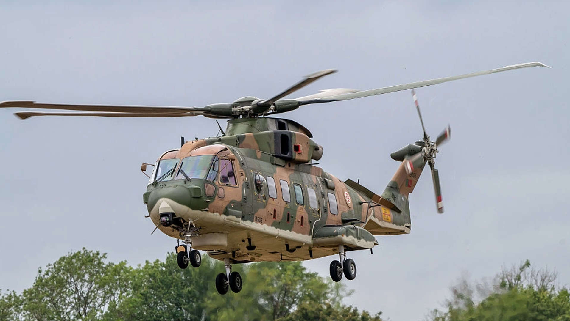A Merlin MK4 helicopter is pictured in flight. It has khaki green and tan-beige camouflage print covering the whole outside, with six windows along the main body and two large front windscreen windows. The background is occupied by grey clouds and tree greenery along the bottom. 