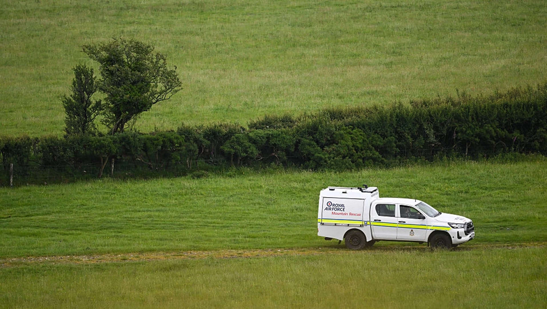 A royal air force truck vehicle drives along in a field in Devon. The field is the site of the crash site. the vehicle is white with the official blue red and white branding of the British royal navy on the side of the back compartment, with a neon yellow stripe across the whole middle of the vehicle. Grass occupies the entire background, with a strip of bush and a single tree across the entre middle, breaking up the image. 