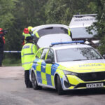 There are multiple police and emergency service vehicles lined up along the left on the image, getting further away from the camera and becoming obscured to the camera by shrubbery and trees. A police officer is walking along a path beside the vehicles towards the camera. Police tape can be seen in the back, cutting off the background. The foreground is occupied mainly by the path/country road and the first police car in the line. The background is occupied mainly by greenery, and several emergency personnel in high vis.
