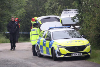 There are multiple police and emergency service vehicles lined up along the left on the image, getting further away from the camera and becoming obscured to the camera by shrubbery and trees. A police officer is walking along a path beside the vehicles towards the camera. Police tape can be seen in the back, cutting off the background. The foreground is occupied mainly by the path/country road and the first police car in the line. The background is occupied mainly by greenery, and several emergency personnel in high vis.