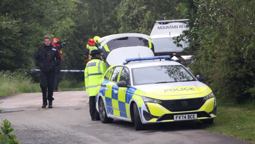 There are multiple police and emergency service vehicles lined up along the left on the image, getting further away from the camera and becoming obscured to the camera by shrubbery and trees. A police officer is walking along a path beside the vehicles towards the camera. Police tape can be seen in the back, cutting off the background. The foreground is occupied mainly by the path/country road and the first police car in the line. The background is occupied mainly by greenery, and several emergency personnel in high vis.