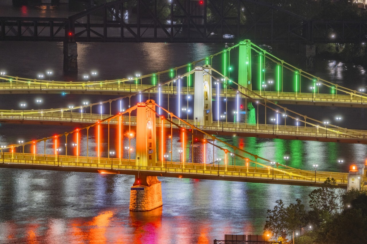 Image shows the Three Sisters bridge in Pittsburgh, lit up the colours orange, white and green at night time