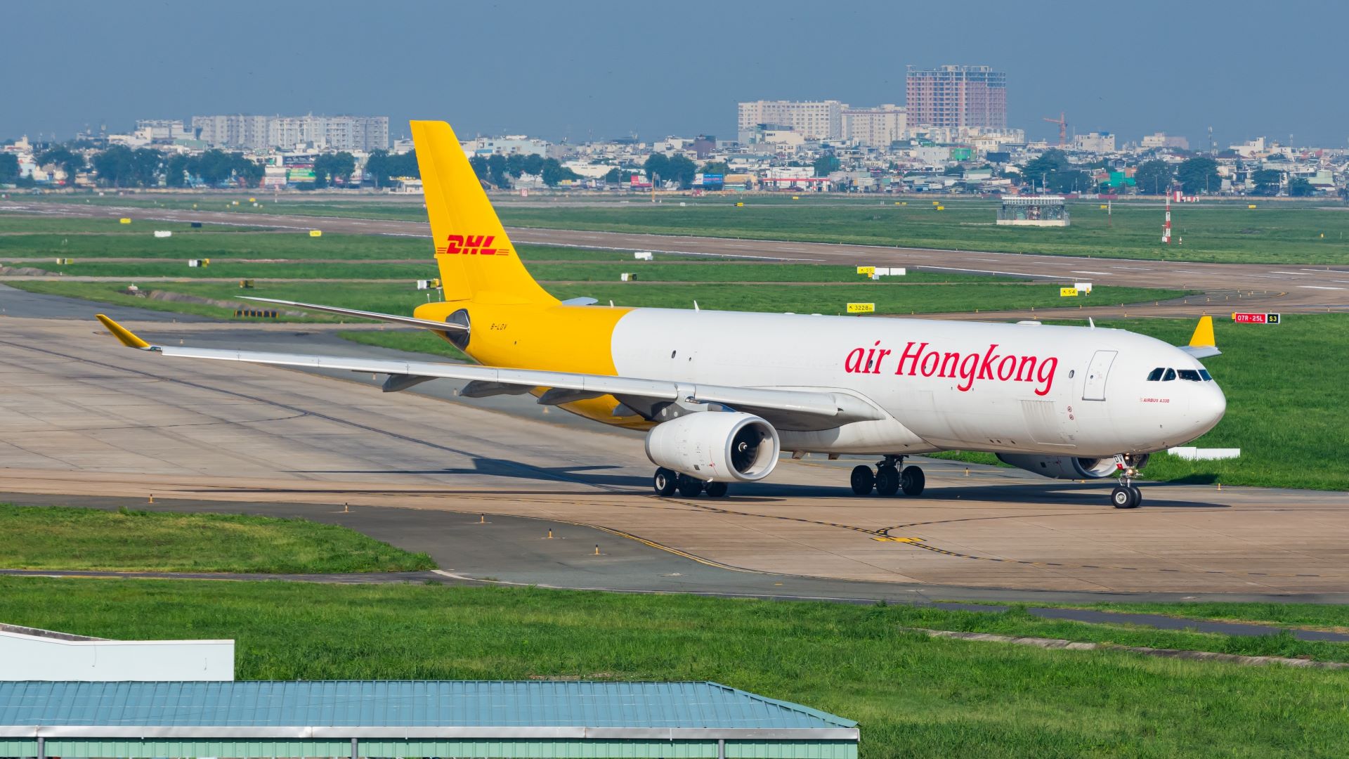 An Air Hong Kong Airbus A330 aircraft on the runway before takeoff. Orange, red and white livery. Blue sky and buildings seen in the background, grass in the foreground.