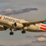 An American Airlines aircraft is in flight, with its nose positioned upper centre left and tail positioned lower centre right. The aircraft is mostly white and has red and blue branding along the middle of the body and on the tail. The sky surrounding the plane is cloudy but light and pale blue