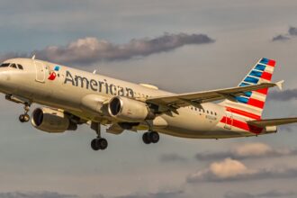 An American Airlines aircraft is in flight, with its nose positioned upper centre left and tail positioned lower centre right. The aircraft is mostly white and has red and blue branding along the middle of the body and on the tail. The sky surrounding the plane is cloudy but light and pale blue