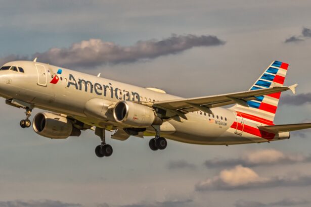 An American Airlines aircraft is in flight, with its nose positioned upper centre left and tail positioned lower centre right. The aircraft is mostly white and has red and blue branding along the middle of the body and on the tail. The sky surrounding the plane is cloudy but light and pale blue