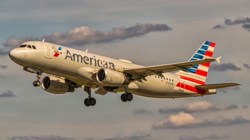 An American Airlines aircraft is in flight, with its nose positioned upper centre left and tail positioned lower centre right. The aircraft is mostly white and has red and blue branding along the middle of the body and on the tail. The sky surrounding the plane is cloudy but light and pale blue