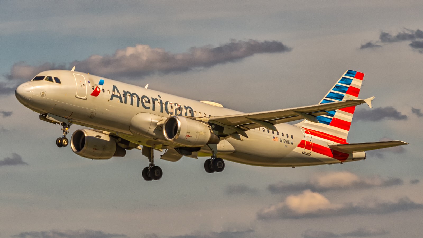 An American Airlines aircraft is in flight, with its nose positioned upper centre left and tail positioned lower centre right. The aircraft is mostly white and has red and blue branding along the middle of the body and on the tail. The sky surrounding the plane is cloudy but light and pale blue