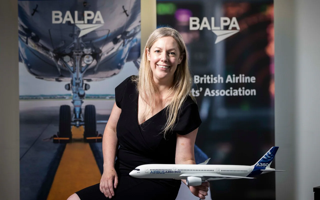 Amy Leversidge seated beside a display holding a model Airbus A350-900 aircraft, positioned in front of banners showing the BALPA logo and an image of an airplane landing gear.