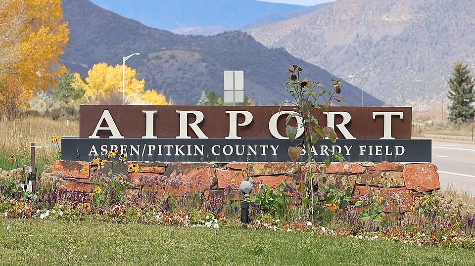 A sign of the Aspen/Pitkin County Airport. Grass in the foreground with mountains and trees seen in the background.