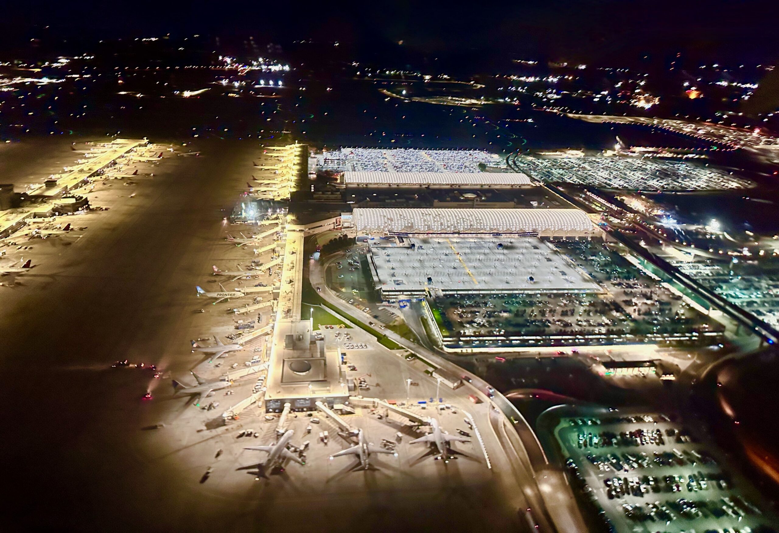 Aerial night-time view of a large, brightly lit airport terminal complex, seen from above with aircraft parked at multiple gates along illuminated concourses. Several wide‑body jets are positioned on the apron in the foreground, with service vehicles and ground equipment clustered around them. The terminal buildings feature long, curved roofs glowing under floodlights, while adjacent multi‑storey car parks are filled with rows of vehicles. Taxiways and runways extend into the distance, marked by lines of blue and green lights, and surrounding roads carry streams of traffic through a dark landscape dotted with city lights.