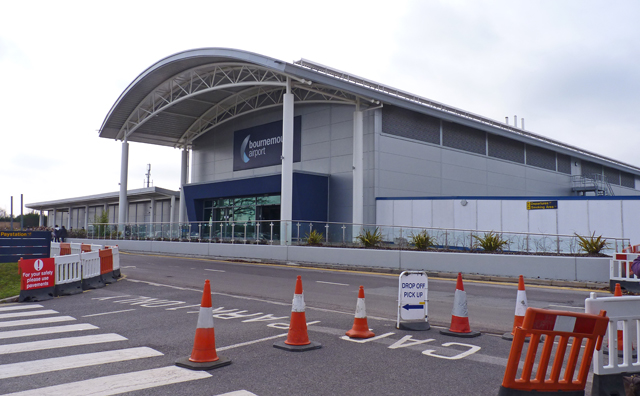 The main terminal building at Bournemouth Airport, a modern grey structure with a curved roof and a blue entrance façade, is pictured behind a road lined with orange traffic cones, pedestrian crossing markings and a temporary “Drop Off Pick Up” sign in the foreground under overcast skies.