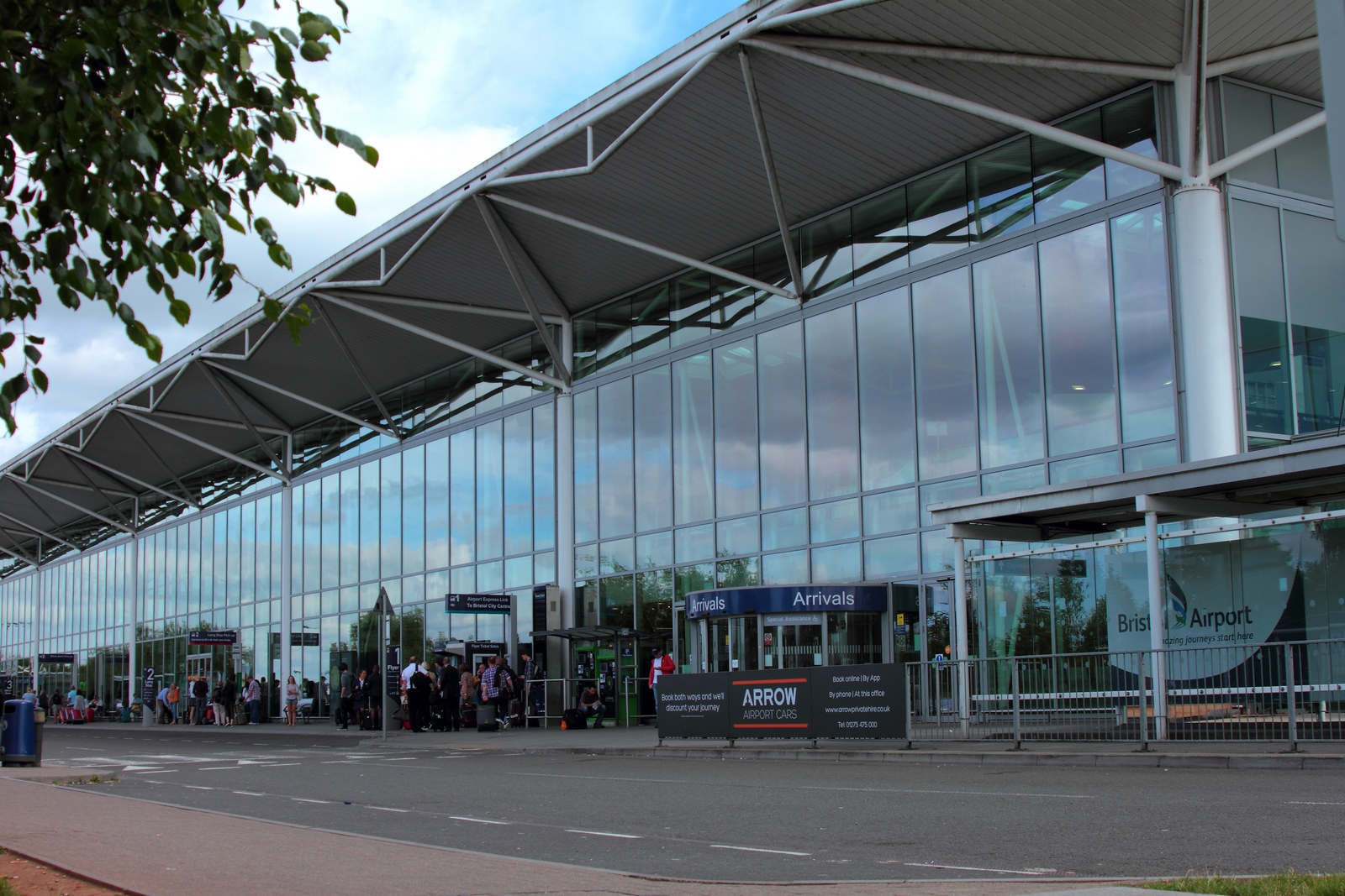 Outside the front entrance for arrivals at Bristol Airport. The entire front of the building is made of glass, with an outside overhang roof extending out from the top. People walk along the pavement around and towards the entrance. A bus stop can be see directly outside on the far right. The image is taken from the pavement across the road from the entrance at an angle, with the building occupying the top right corner and lower mid left. In the immediate foreground is a bus stop drop off point. In the left hand corner is blue sky dotted with clouds and some foliage from an unseen tree. 