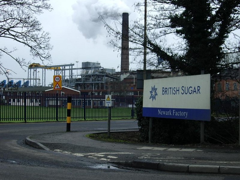 Taken from across the road, the image shows a large brick factory with a tall chimney puffing smoke . In the foreground, on the centre right angled towards the camera, a white sign can be seen reading 'British Sugar' in blue with a logo. Beneath this, the sign's colour scheme reverses and the words 'Newark Factory' are written in white on a blue strip at the bottom. It appears to be a cloudy and wet day. Road signs, a gate around the factory, and roadside bollards can be seen in the image.