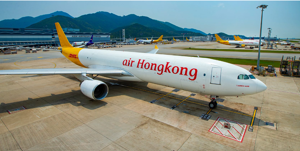 An Air Hong Kong A330 aircraft sat on the apron with white, red and orange livery. Aircraft seen behind, and there are mountains in the background.