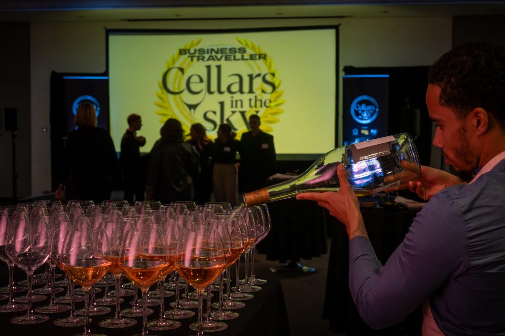 a man pouring a bottle of wine into numerous glasses on a table, with the business traveller cellars in the sky logo projected in the background